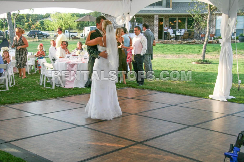 A bride and groom dancing together on a wooden dance floor at an outdoor wedding reception, surrounded by guests at tables celebrating in a festive atmosphere.