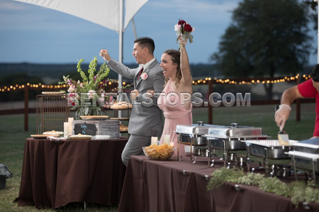 A joyful couple celebrating at an outdoor event, with the woman in a light pink dress holding flowers and the man in a gray suit raising his arms. Tables with food and flowers are set up in the background.