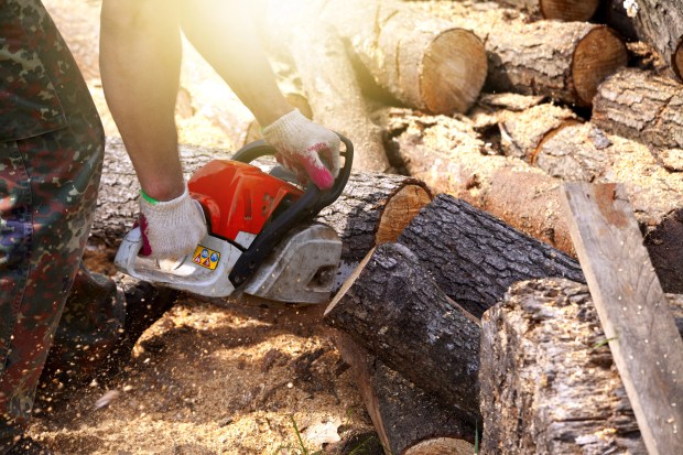 A person using a chainsaw to cut logs in a woodpile, wearing gloves and standing on sawdust-covered ground.