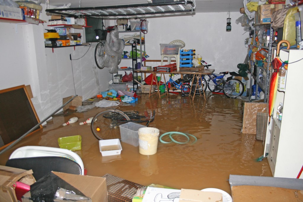 A flooded garage filled with water, with bicycles, a wooden table, and various items scattered around, including boxes and plastic containers.