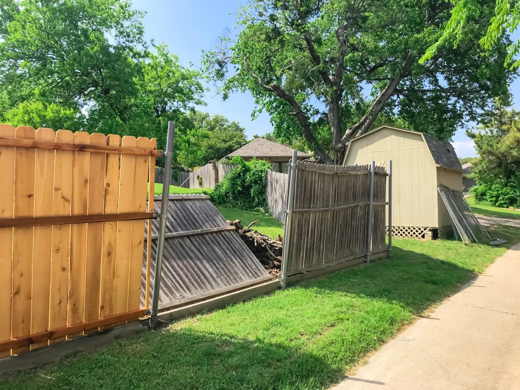 A wooden fence with a broken section leaning to the side, revealing a pile of debris behind it. Lush green trees and a small shed are visible in the background.