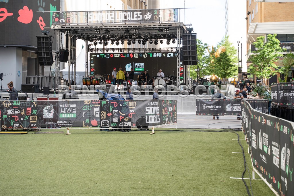 An outdoor stage setup for a live music event, featuring a large sound system and colorful banners with branding. The area has green grass, with people preparing for the event in the background.
