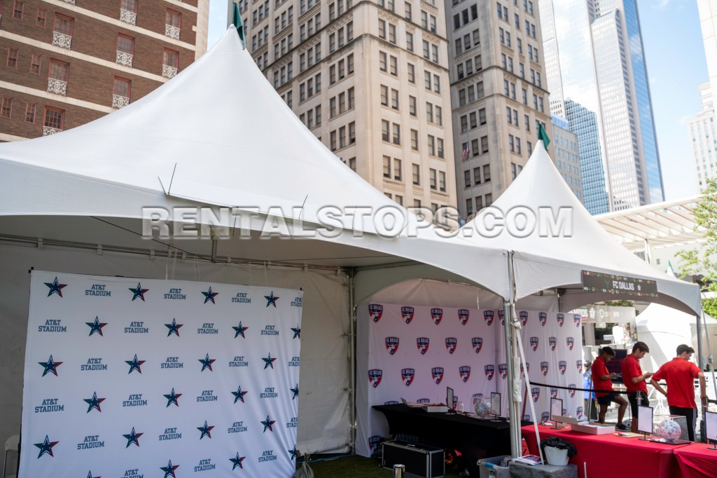 Outdoor event setup featuring large tents with branding for AT&T Stadium and FC Dallas. Background includes tall buildings and a clear sky.