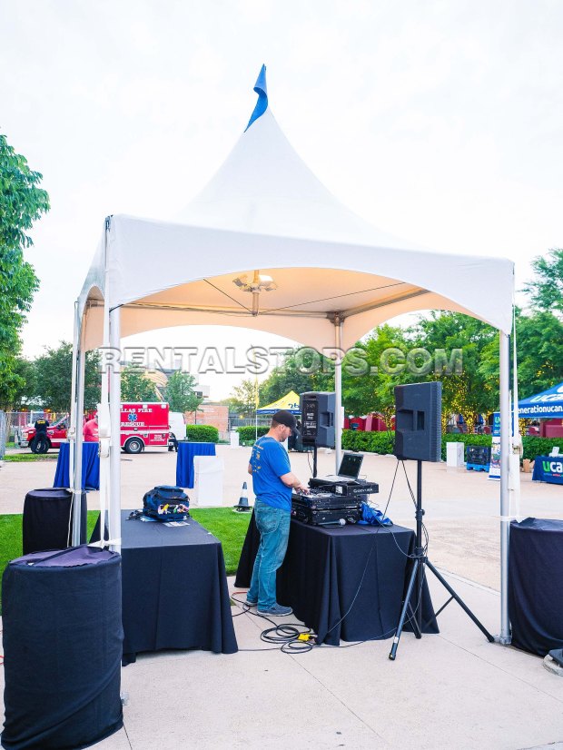 A DJ performing under a white tent at an outdoor event, with sound equipment and tables.