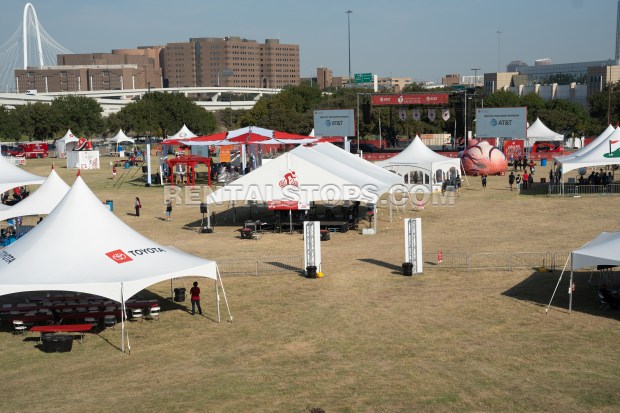 A large outdoor event scene with multiple white tents set up on a grassy area, surrounded by trees and city buildings in the background. Some tents have logos visible, and a stage with screens is set up in the distance.