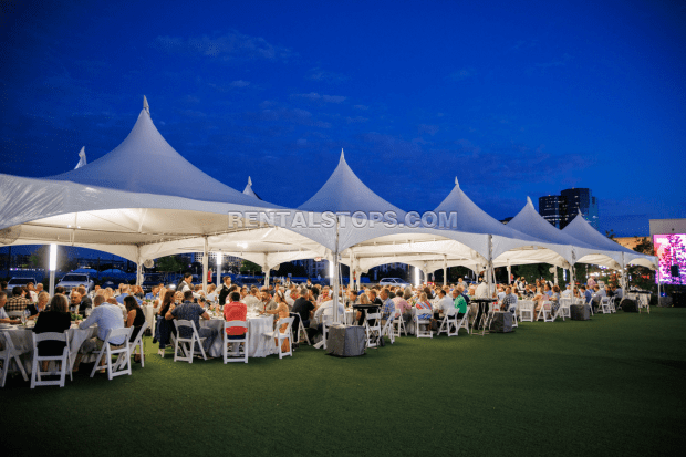 An outdoor dining event under large white tents at dusk, with numerous guests seated at long tables.