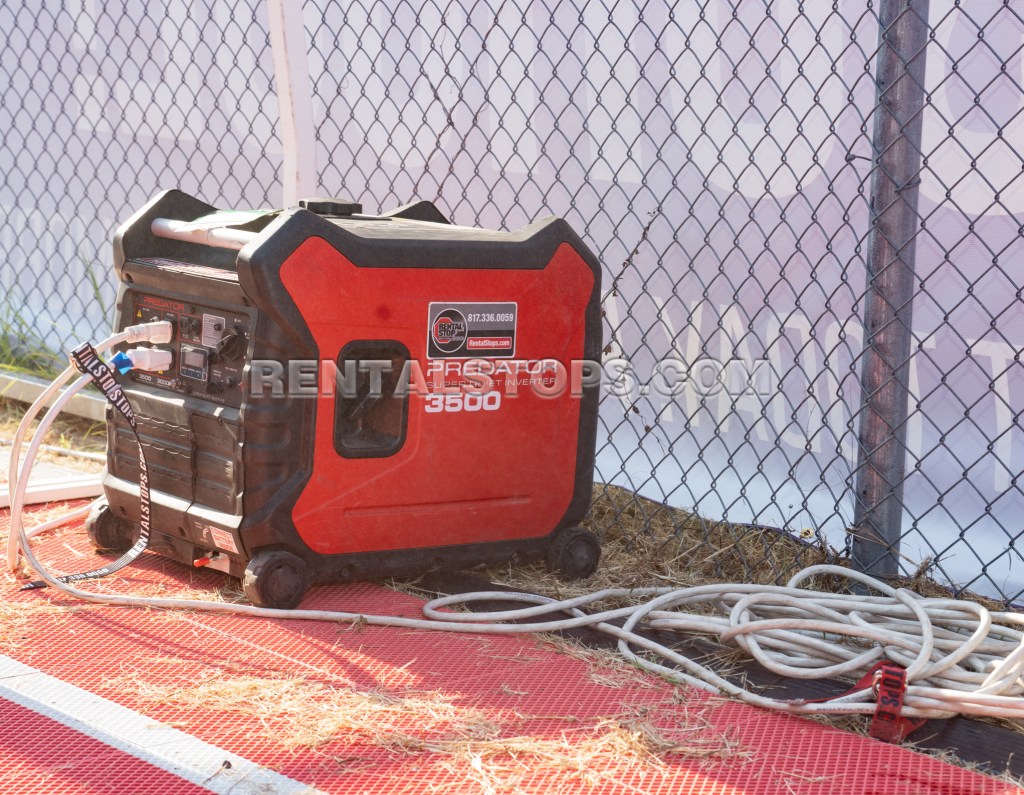 A black and red portable generator placed on a red surface, with power cables connected, beside a chain-link fence.