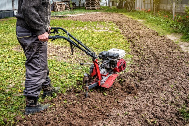 A person using a red and black tiller to cultivate soil in a garden, wearing gray pants and boots, with green foliage in the background.
